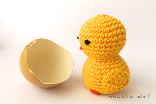 Un poussin amigurumi en crochet à côté d’un morceau de coquille d’œuf